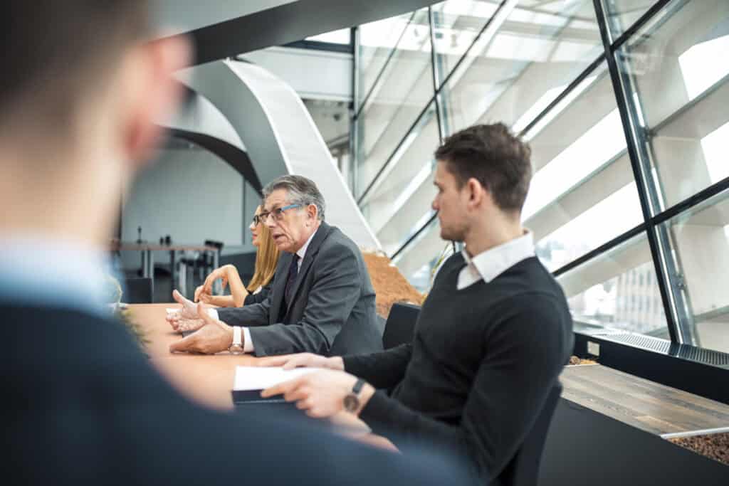 People in business attire sit at a table in a modern office with large windows, engaged in discussion.