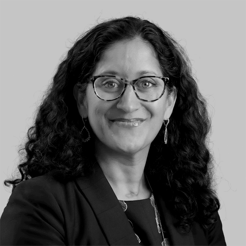 Woman with curly hair and glasses, wearing a blazer and earrings, smiling at the camera against a plain background.