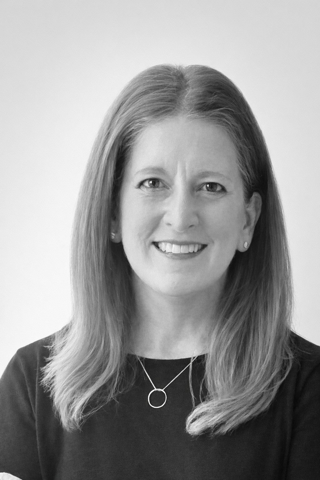 Smiling woman with straight, shoulder-length hair wearing a dark top and a circular pendant necklace.