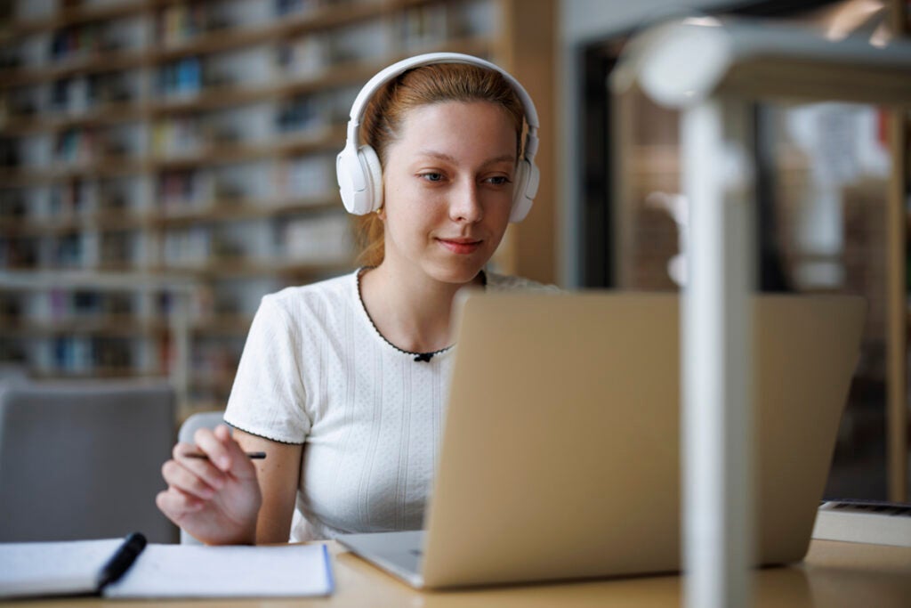 Young woman wearing headphones studies at a library table with a laptop, notebook, and pen.